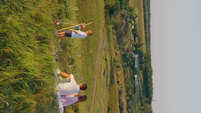 Vertical Screen: Family spending leisure in meadow at sunset, parents on ground and watching little son drawing picture on easel, country houses on background. Creative child painting in open air