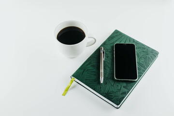 Flat lay of, cell phone, pen, and green notebook on white background with cup of coffee