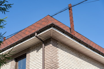 Brown roof of house covered with flexible bitumen motleysoft shingles under blue sky on sunny day. Cottage roof with metallic guttering system, guttering and drainage pipe exterior