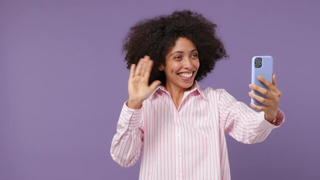 Fun young black woman 20s wears pink shirt get video call using mobile cell phone talk conducting pleasant conversation greet with hand isolated on plain pastel light purple background studio portrait