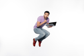 shocked man in jeans and purple t-shirt levitating while holding laptop on white.