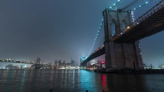 Zooming in timelapse of the illuminated at night Manhattan and Brooklyn bridge on the left that cross the East River in New York City, USA. Vessels sail along the illuminated river, skyscrapers