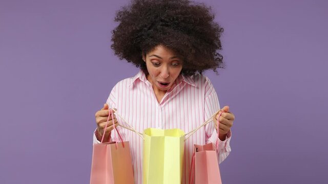 Happy shocked amazed young black woman 20s years old wears pink shirt holding looking into package bags with purchases after shopping isolated on plain pastel light purple background studio portrait