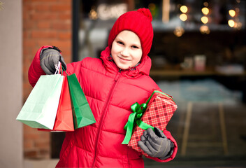 Boy kid in winter clothes standing outside near by shop market, holding gift box and paper bags. Christmas and New Year shopping concept.