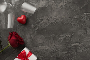 Top view photo of saint valentine's day decorations two wineglasses white giftbox with red bow small heart and red rose on isolated textured dark grey concrete background with copyspace