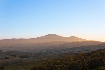 Photo of a Tuscan countryside panorama during sunset. In the foreground the cultivated fields and a wood, in the background some houses, the mountains and the sun's rays