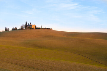 Fototapeta na wymiar Hills with plowed fields in the Italian countryside in Val D'Orcia, photographed at sunset. Warm light, with a cottage and trees in the background. Relaxing landscape with lines formed by plow furrows