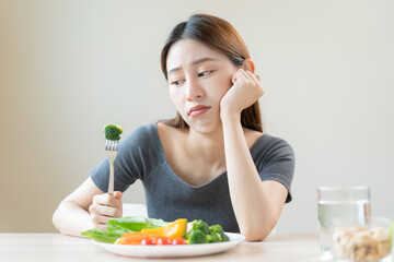 Obraz na płótnie Canvas Diet in bored, unhappy beautiful asian young woman, girl on dieting, holding fork at broccoli in salad plate, dislike or tired with eat fresh vegetables. Nutrition of clean, healthy food good taste.