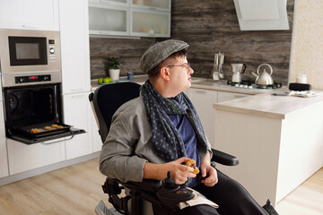 Man in casualwear eating homemade cookie while sitting in wheelchair against open electric oven...