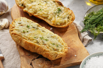 Two halves of garlic and butter bread - baguette on a wooden board, sea salt, pepper, dill and garlic cloves on a grey concrete background, top view