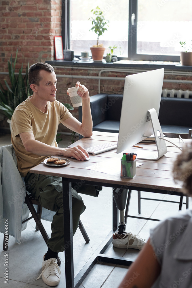 Poster young serious businessman with leg prosthesis sitting by workplace in front of computer monitor and 