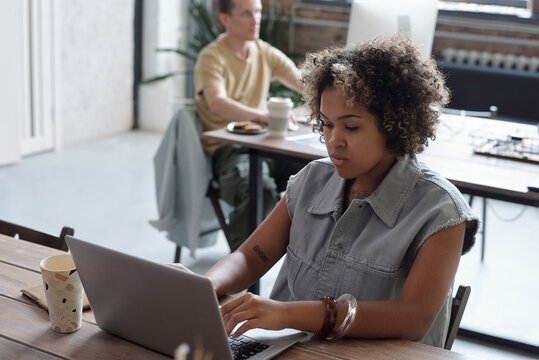 Young biracial businesswoman pressing keys of laptop keypad while sitting by table against male colleague in openspace office