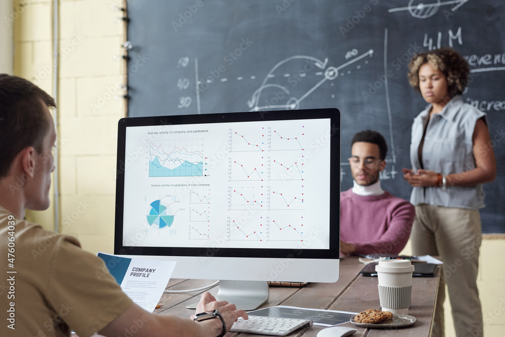 Poster Young serious broker analyzing financial graphs on computer screen while sitting by table in front of two intercultural colleagues