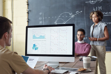 Young serious broker analyzing financial graphs on computer screen while sitting by table in front of two intercultural colleagues