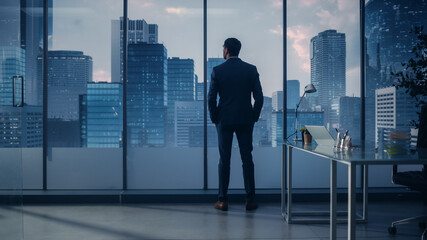 Fototapeta na wymiar Confident Young Businessman in a Perfect Tailored Suit Standing in His Modern Office Looking out of the Window on Big City with Skyscrapers. Successful Finance Manager Planning Project Strategy.