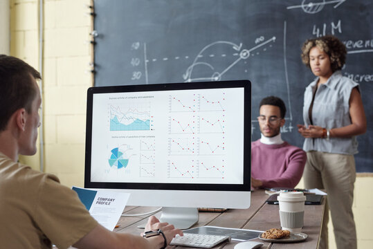 Young serious broker analyzing financial graphs on computer screen while sitting by table in front of two intercultural colleagues