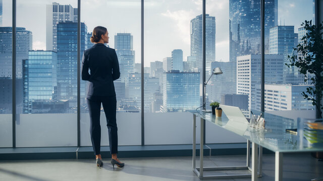 Successful Businesswoman In Stylish Suit Looking Out Of The Window At Big City In Downtown Area. Confident Female CEO Working On Investment Opportunity. Real Estate Agent Planning A Development Deal.