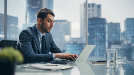 Happy Businessman in a Suit Sitting at a Desk in Modern Office, Using Laptop Computer, Next to Window with Big City with Skyscrapers View. Successful Finance Manager Planning Work Projects.