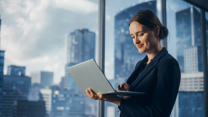 Successful Businesswoman in Stylish Dress Working on Laptop, Standing Next to Window in Big City....