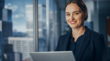 Successful Businesswoman in Stylish Dress Working on Laptop, Standing Next to Window in Big City....