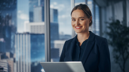 Successful Businesswoman in Stylish Dress Working on Laptop, Standing Next to Window in Big City. Confident Female CEO Analyze Financial Projects. Manager at Work Planning Marketing Campaign.