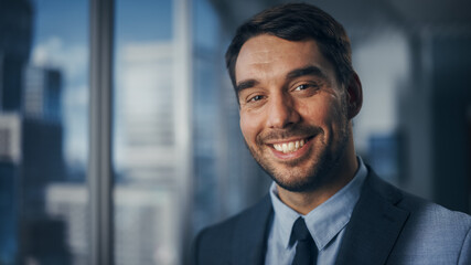 Close Up Portrait of Handsome Caucasian Businessman in Stylish Black Suit Posing Next to Window in Big City Office with Skyscrapers. Confident Male CEO Smiling. Successful Masculine Business Manager.