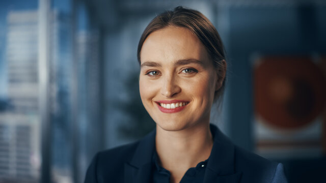 Close Up Beautiful Portrait Of A Businesswoman In Stylish Black Suit Posing Next To Window In Big City Office With Skyscrapers. Confident Female CEO Smiling. Successful Diverse Business Manager.