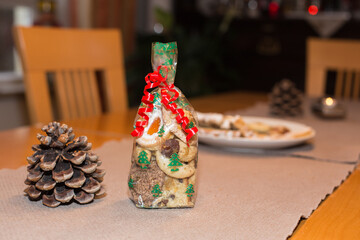 Seasonal homemade advent or christmas cookies in a transparent bag gift wrapped with curled red ribbon placed on a table which is decorated with a fir cone, bokeh lights, blurred background