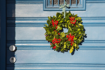 Decorative Advent wreath or crown with red berries bonded with holly or ilex branches and foliage and fixed at the window of a house entrance door with a fabric ribbon