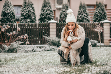 Beautiful woman in warm clothes and white knitted hat kisses her beloved dog in snow-covered garden. Cavalier King Charles spaniel. Wonderful moments Love and care. Snow is falling.