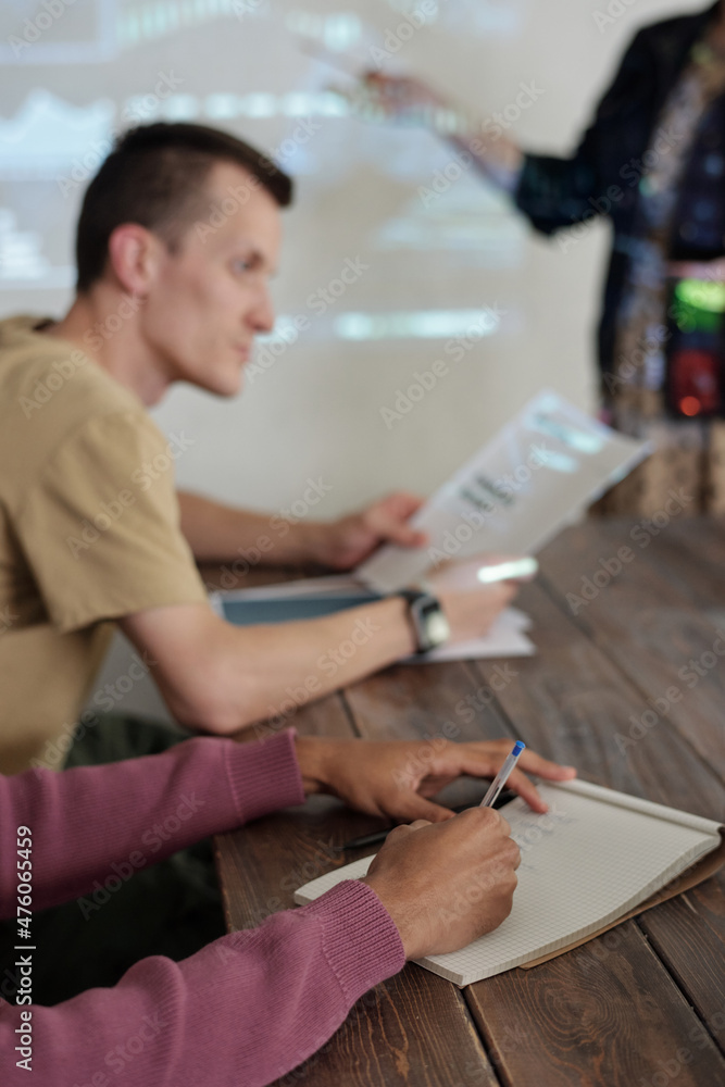 Poster Hands of young biracial businessman making notes in notepad while sitting by table during presentation at seminar or business training