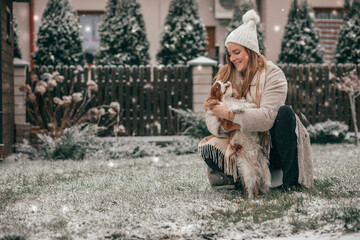 Charming young woman in white knitted hat is playing in snowy garden with dog. Cavalier King Charles spaniel. Winter landscape. New Year's card. Pets. Wonderful moments . Lot of happiness.