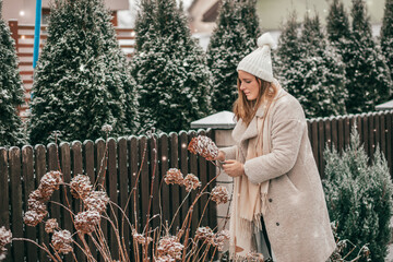 Young woman in winter clothes and knitted white hat with scarf in the winter garden chooses snow -...