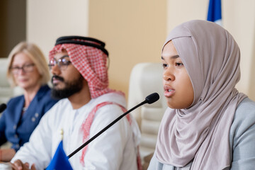 Young contemporary female delegate in hijab making speech while sitting against Arabian and...