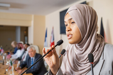 Pretty young Muslim woman in hijab speaking in microphone while standing in front of audience...