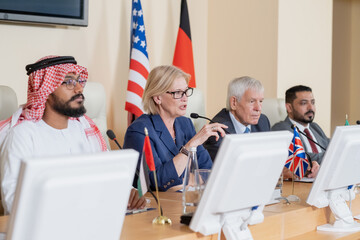 Blond mature female delegate making speech for audience while sitting by table between several...