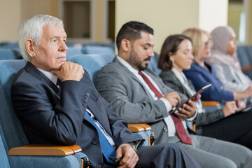 Senior businessman or politician in formalwear listening to speaker at conference while sitting in...
