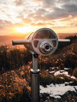 Close up image of a binoculars (telescope) on the hill in nature with beautiful sunset. Photo of outdoor tourist telescope in winter forest with city and hills on background - dramatic cloudy sky.