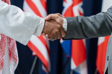 Two contemporary male delegates or political leaders shaking hands against flags while standing in...