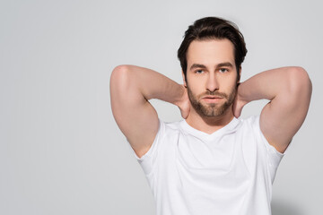 brunette bearded man in white t-shirt holding hands behind neck while looking at camera on grey.