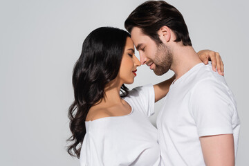 side view of brunette woman embracing man while standing face to face with closed eyes isolated on grey.