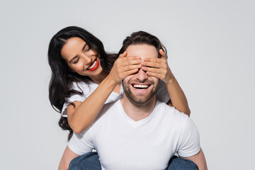 Naklejka premium joyful woman covering eyes of smiling man while playing guess who game isolated on grey.