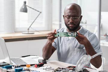 Young serious African man in eyeglasses and uniform looking at circuit board while going to repair...