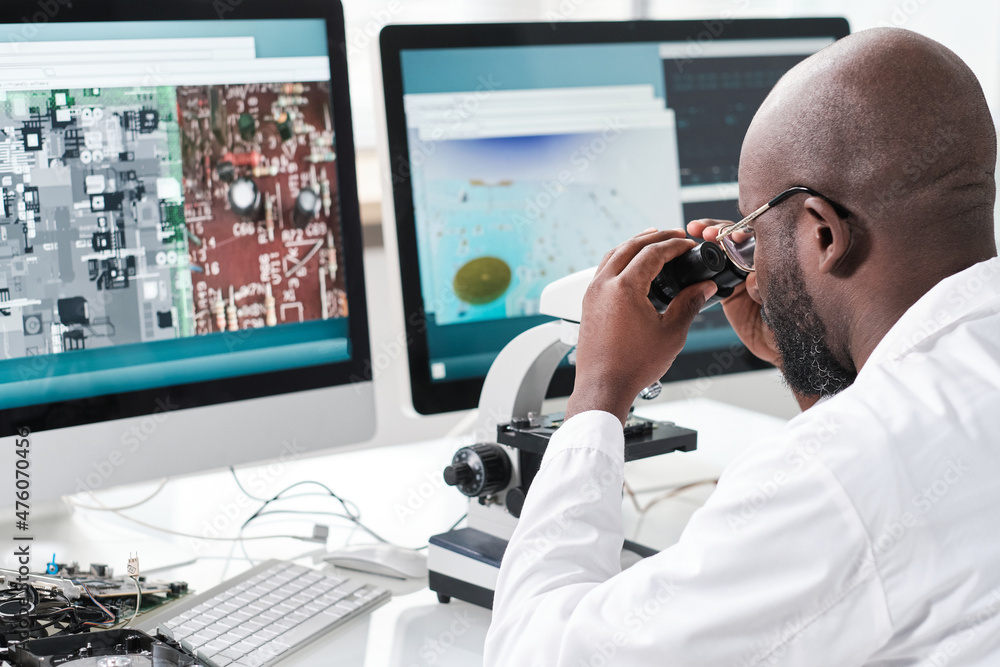 Poster Young man of African ethnicity looking in microscope while sitting in front of computer screens with magnified microchip