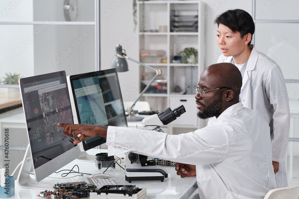 Poster Young confident scientist pointing at magnified microchip scheme on computer screen during discussion with female colleague