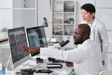 Young confident scientist pointing at magnified microchip scheme on computer screen during discussion with female colleague