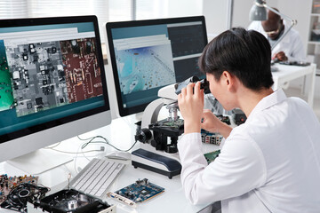 Young Asian female researcher in whitecoat looking in microscope while sitting by desk in front of...