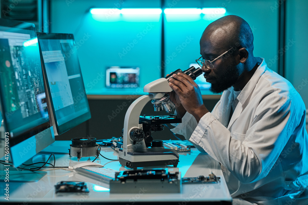 Poster Young serious male researcher in whitecoat looking in microscope while studying microprocessor in front of computer monitors