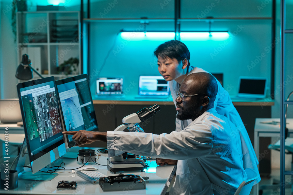 Poster Two young intercultural professionals in whitecoats discussing microchip scheme while one of them pointing at computer screen