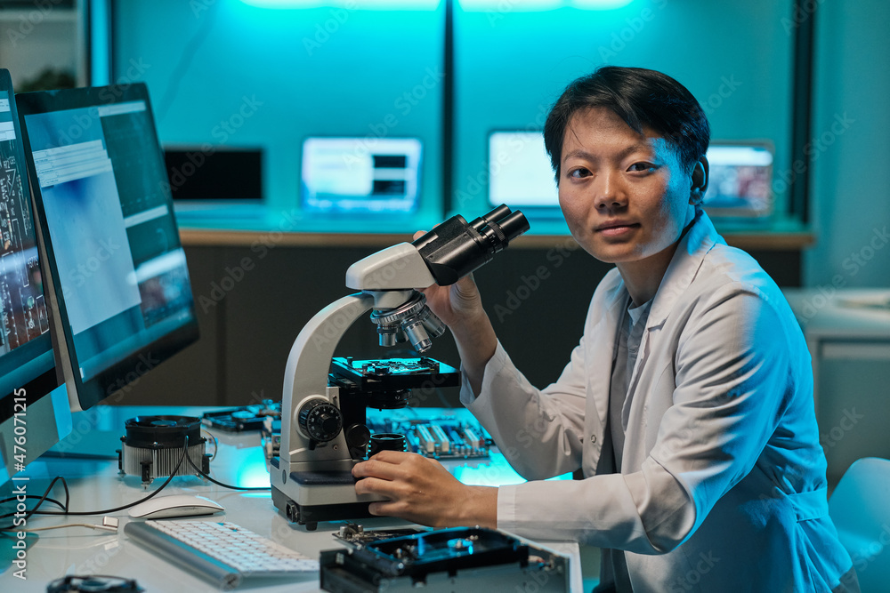 Poster young asian researcher in whitecoat working with microscope and looking at you while sitting by work